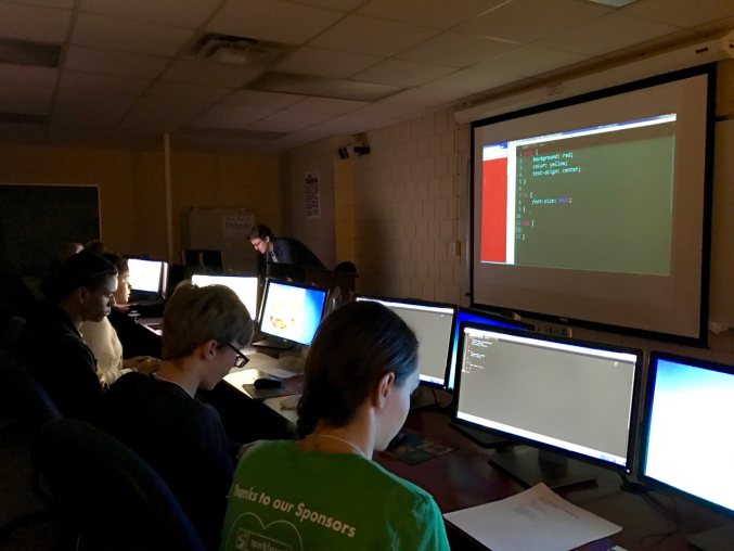 Young people working at a series of computers in front of a projector screen in a dark room