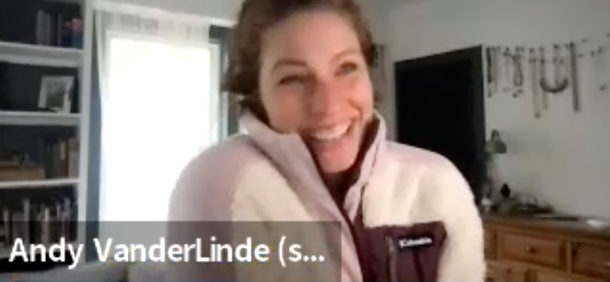 A woman in a light pink pullover smiling at the Zoom camera. A bookshelf and window are in the background.